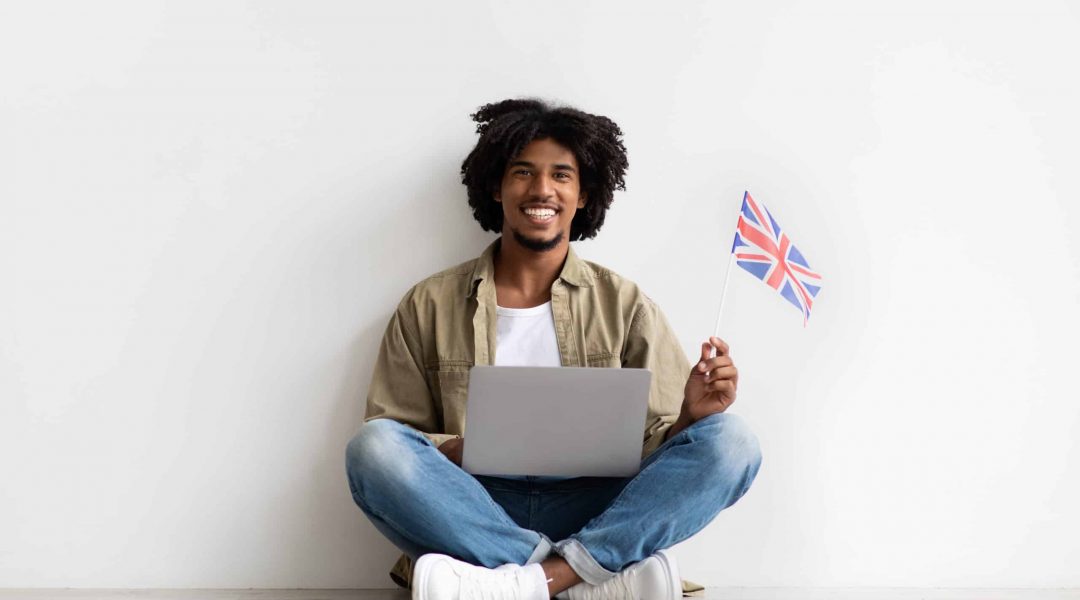 Smiling African American Guy Sitting On Floor With Laptop And Great Britain Flag, Happy Young Black Man With Computer Recommending Language Courses, Advertising Online Education Website, Copy Space